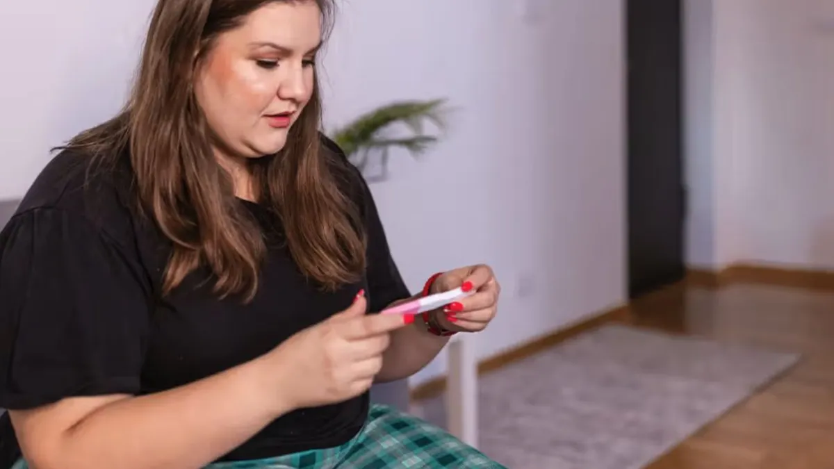 A woman with dark brown hair wearing a black t shirt and green pants looks at a pregnancy test