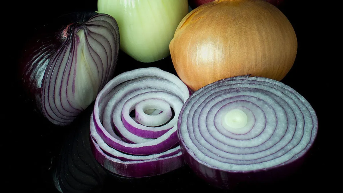 A photograph showing a collection of onions including a whole brown onion, half of a white onion, and several slices of red onions arranged on a reflective black surface