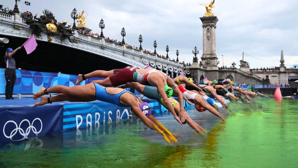 A group of swimmers jump off an Olympic platform underneath a historic bridge into a green river
