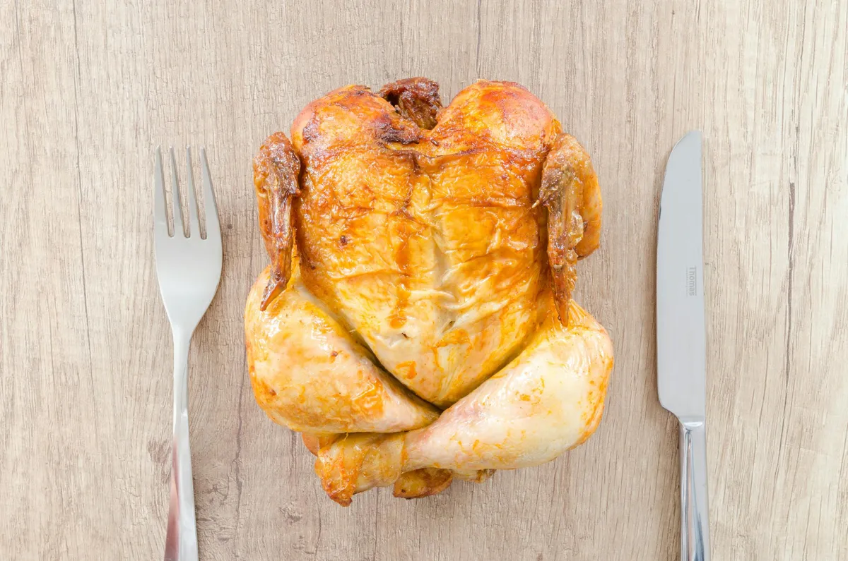 A roasted chicken on a table next to a fork and knife