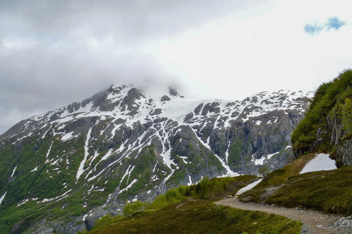 A mountain in Alaska covered in snow and green grass, the top of it covered with clouds.