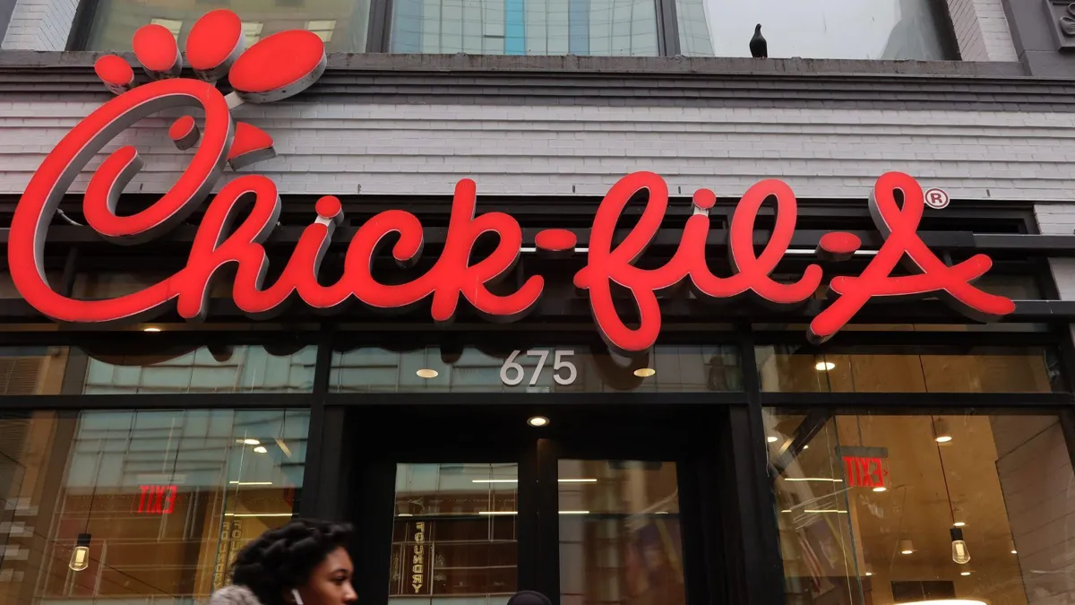 This is the outside of a Chick-fil-A restaurant. The sign is hanging above the restaurant door, and a person is walking past.