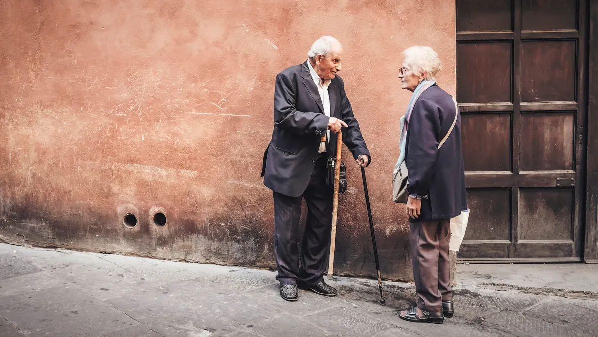 Two elderly people standing next to a lime-washed wall on a cobble road; Researches Unveil the Maximum Age Humans Can Live To
