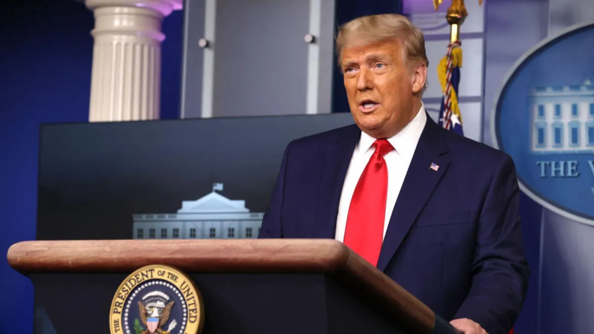 Former President Donald Trump stands at the podium with the Presidential Seal in the White House briefing room. He is wearing a dark suit, white shirt, and red tie. In the background, there is an American flag and a column