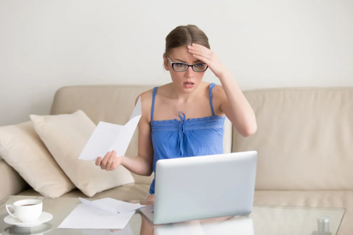 A woman in a blue shirt is pictured with her hand on her head after examining her monthly expenses