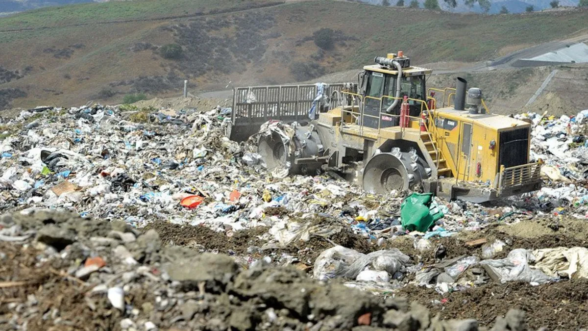 A large yellow landfill compactor vehicle is actively working amidst a vast expanse of waste and debris. The image captures the machine as it compresses and maneuvers over piles of mixed trash, with the rugged terrain of the landfill site visible in the background