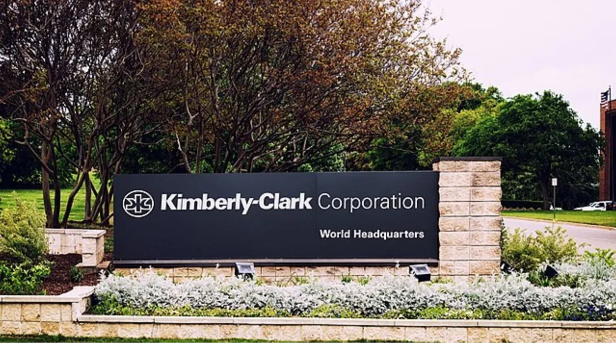 A black sign that says “Kimberly-Clark Corporation World Headquarters”. It is against a brick wall and in front of some trees and a driveway.
