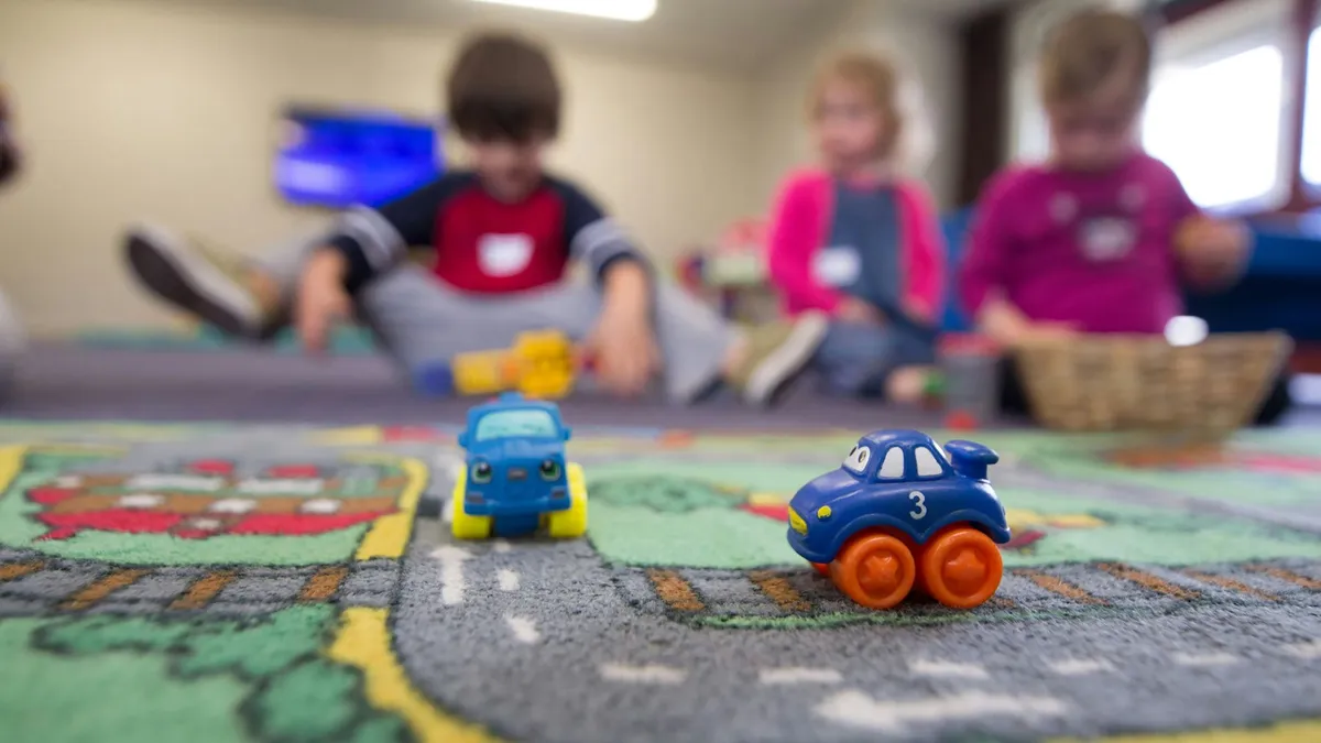 Three kids sit on the ground with two small plastic cars in the foreground