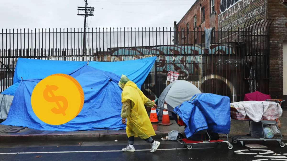 A person wearing a yellow rain slicker pulling a shopping cart full of personal items while walking past a blue tarp