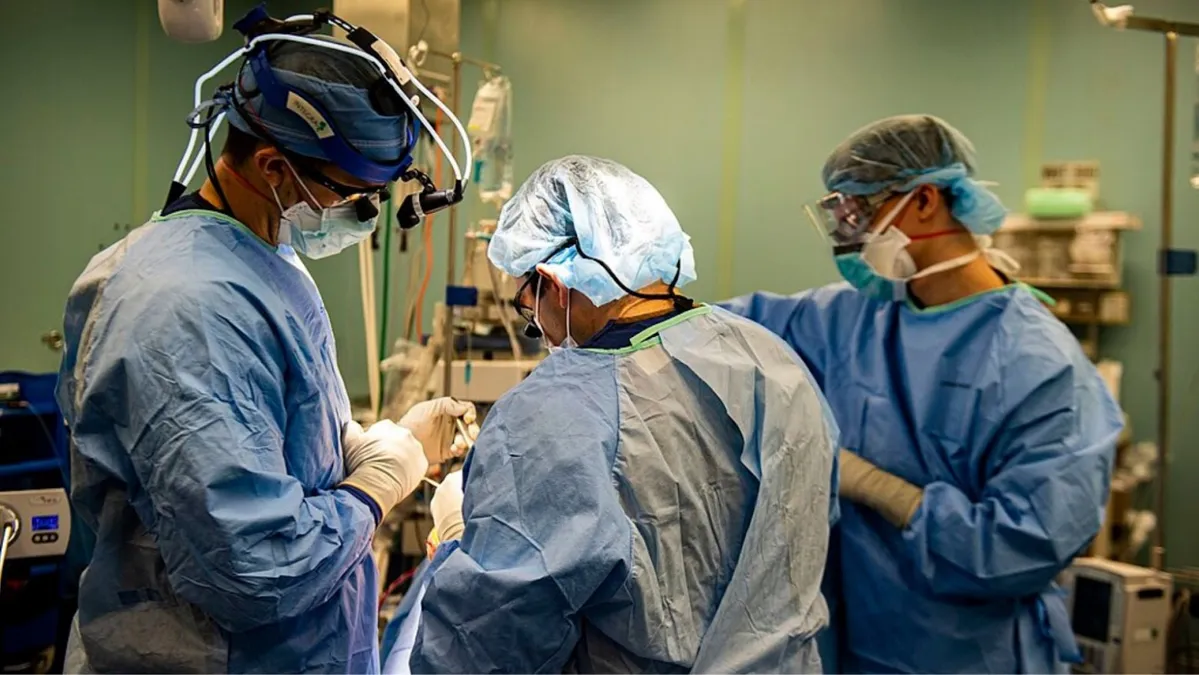 Three doctors wearing PPE and blue scrubs, masks, glasses, and scrub caps perform a surgery