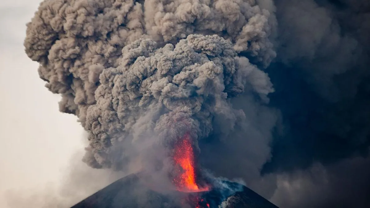 A large plume of black smoke and red lava shoots out of the top of a volcano