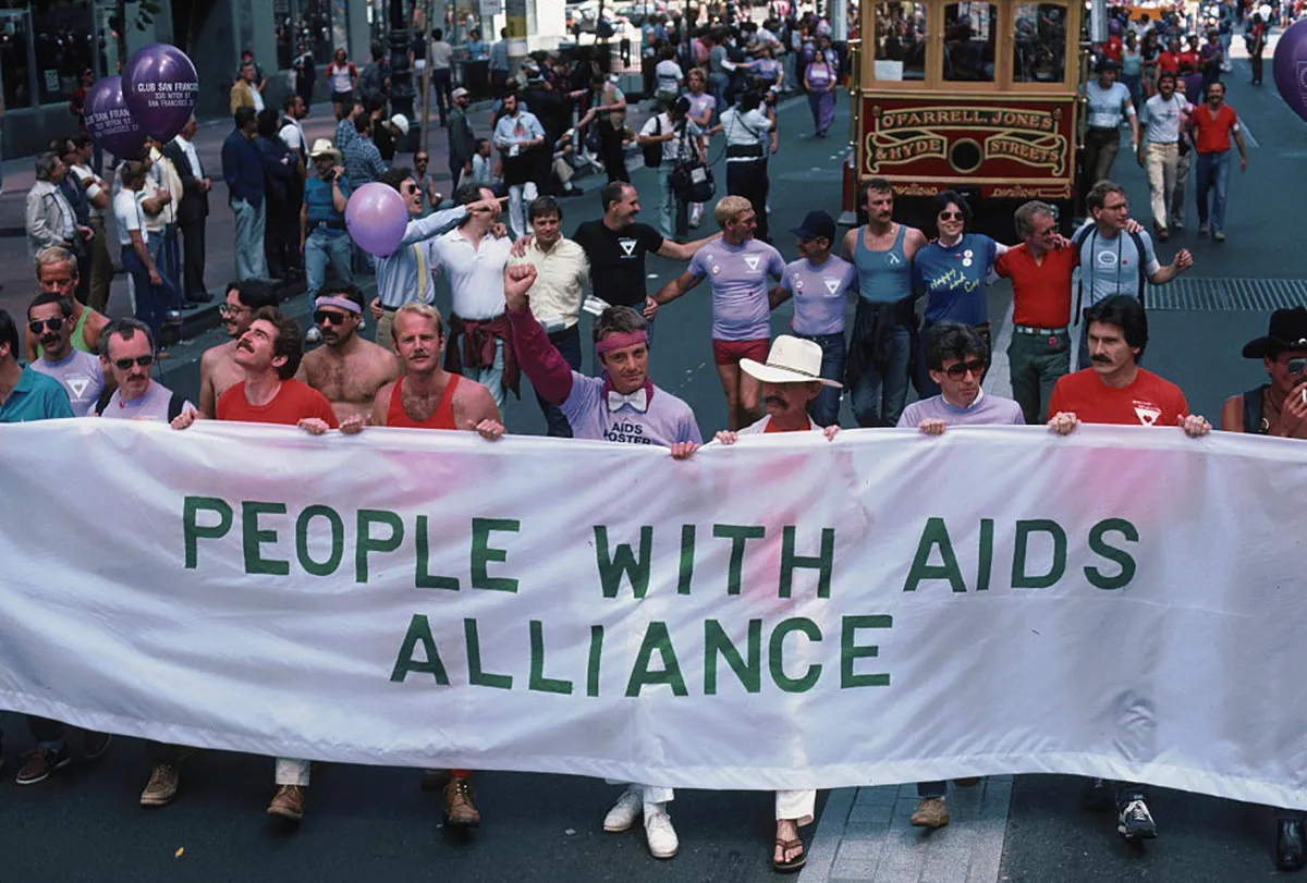 A photograph from the 1980s at an AIDS protest with several men holding a sign that reads “People With Aids Alliance”